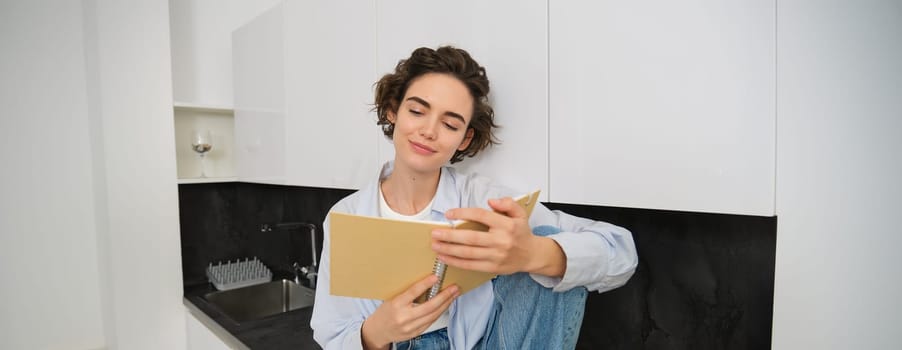 Portrait of smiling young woman reading journal, enjoys comfort at home, holding notebook, looking happy, relaxing indoors.