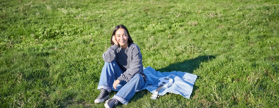 Young asian girl enjoying sunny day outdoors. Happy student having picnic on grass in park, playing ukulele.