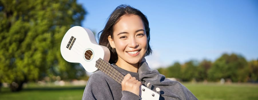 Portrait of beautiful smiling girl with ukulele, asian woman with musical instrument posing outdoors in green park. People and lifestyle