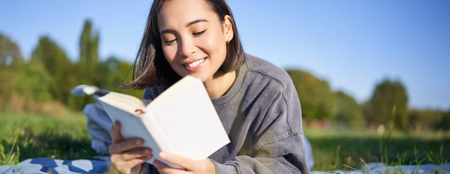 Portrait of beautiful smiling asian girl, reading in park, lying on grass with favourite book. Leisure and people concept.