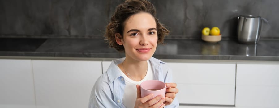 Portrait of smiling, beautiful young woman, drinking coffee in kitchen, morning magic with cuppa tea, looking tenderly.