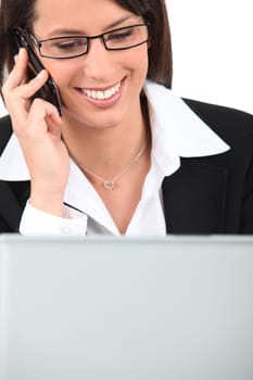 Smiling young woman talking on a cellphone while using a laptop computer