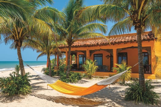 A vibrant hammock hangs between palm trees on the sandy beach beside a colorful coastal villa under clear blue skies.