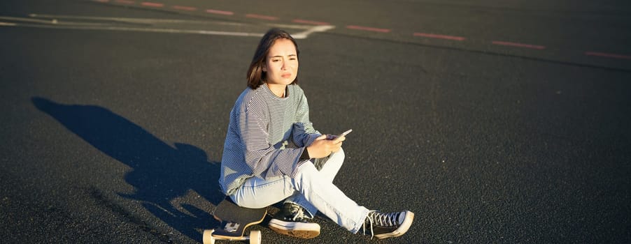 Sad asian teen girl sits on skateboard, looks upset, holds mobile phone and frowns. Copy space