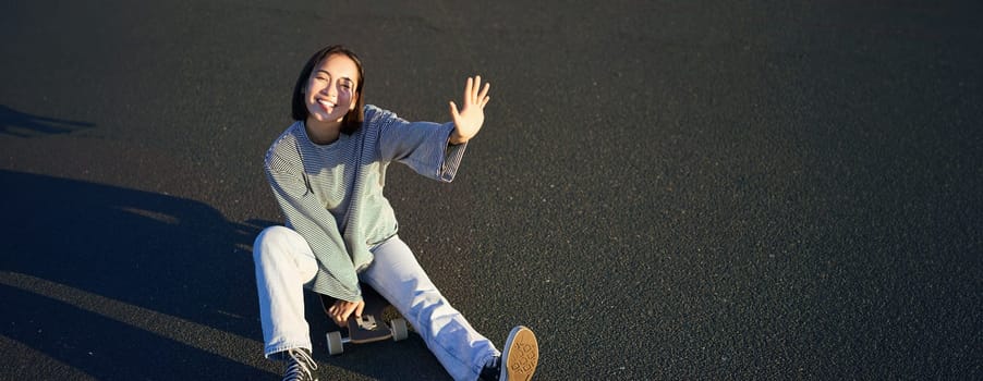 Positive korean girl covers her face from sunlight, sits on skateboard and smiles happily. Copy space