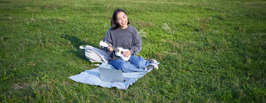 Cute korean girl, musician sits in park, plays ukulele and sings, looks up chords and tutorials on laptop.