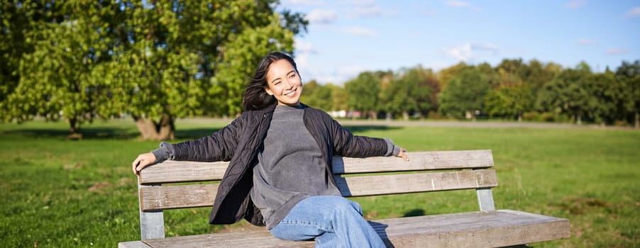 Portrait of young woman in outdoor clothes, sitting on bench relaxed, smiling and enjoying view on green park.