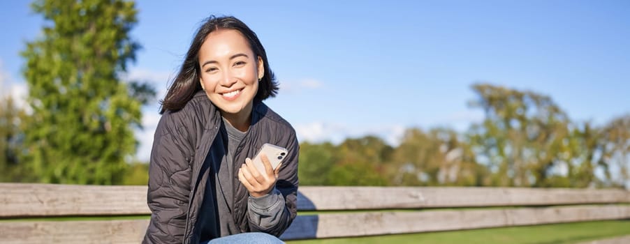 Cute young woman with smartphone in hands, sitting on bench and smiling, using mobile phone, waiting for someone in park.