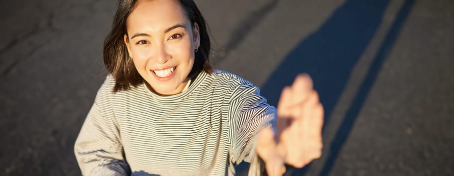 Selfie of beautiful asian girl smiling, taking photo on smartphone while sitting on skateboard outdoors.