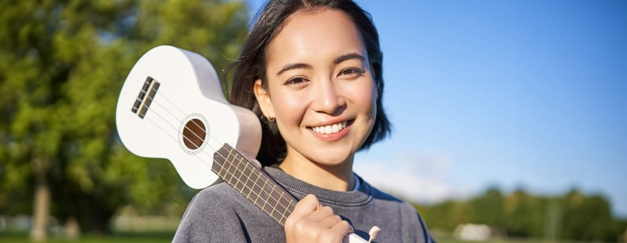 Portrait of beautiful smiling girl with ukulele, asian woman with musical instrument posing outdoors in green park. People and lifestyle