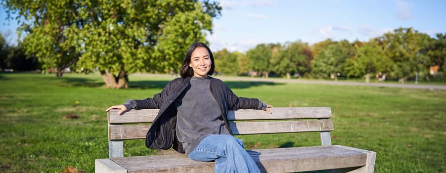 Smiling young asian woman in outdoor jacket, sitting on bench in green sunny park, resting alone, relaxing on fresh air.