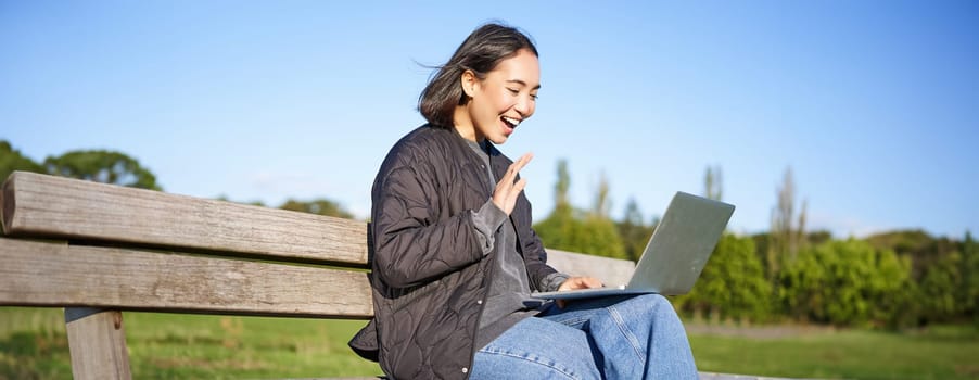 Portrait of young asian woman working remotely from park, sitting on bench with laptop, talking during video chat.