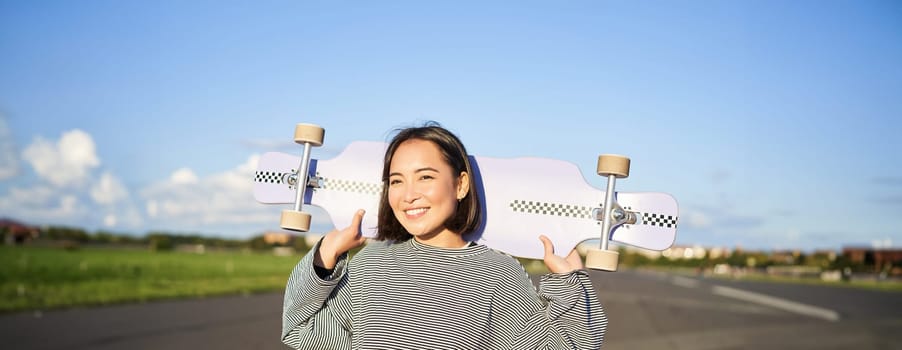 Lifestyle and people. Young asian girl posing with longboard, skating on her cruiser. Smiling woman holding skateboard on shoulders.