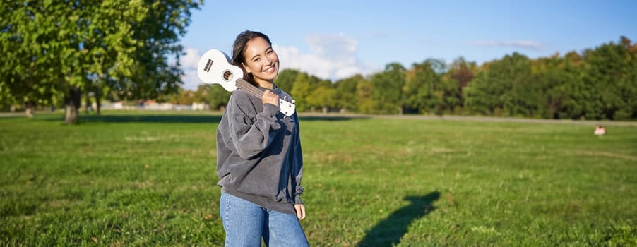 Beautiful korean girl posing with ukulele, young musician playing outdoors in green park on sunny day.