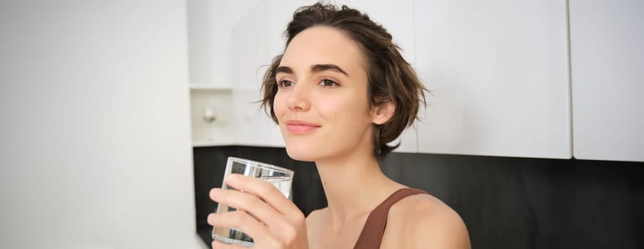 Close up portrait of smiling fitness woman, holding glass of water and lookins away pleased. Girl drinks after workout. Sport and healthy lifestyle concept