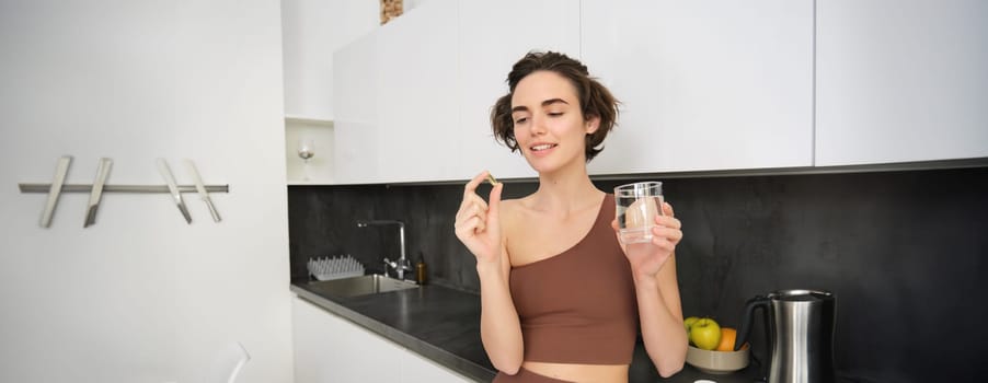 Dietary supplements and healthy lifestyle. Young woman taking vitamin C, D omega-3 with glass of water, standing in activewear, drinking after workout training in her kitchen.