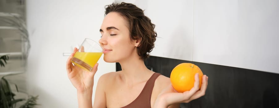 Image of sportswoman, fitness girl holding glass of juice and an orange, smiling, drinking vitamin beverage after workout, standing in her kitchen at home. Healthy lifestyle and sport concept.