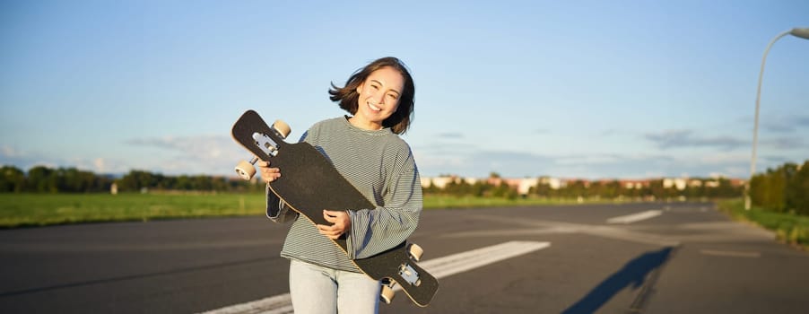 Lifestyle and hobbies. Smiling cute asian girl holding skateboard and walking towards sun on an empty road.