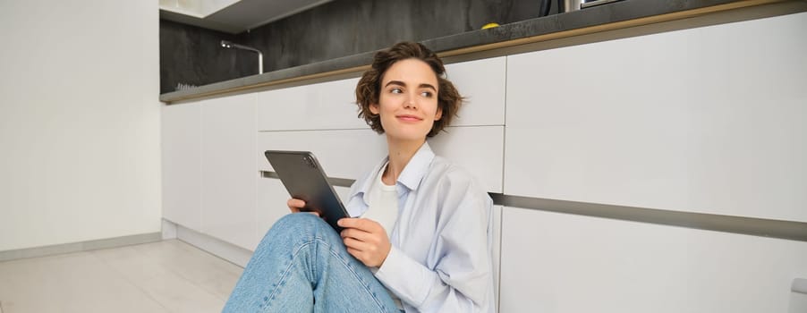Image of happy young woman sits at home on floor, holds digital tablet, does online shopping, reading e-book. Copy space