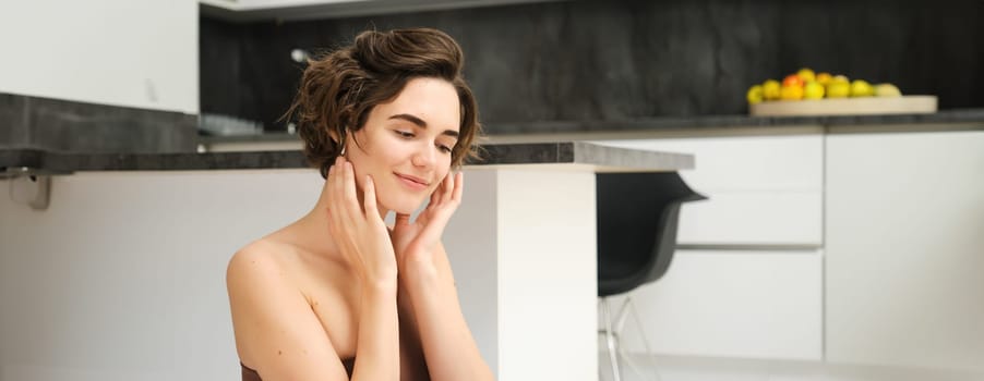 Close up of young smiling brunette woman, fitness girl in kitchen, looks aside, touches her face, listens to mindful app training session in wireless earphones, workout at home.
