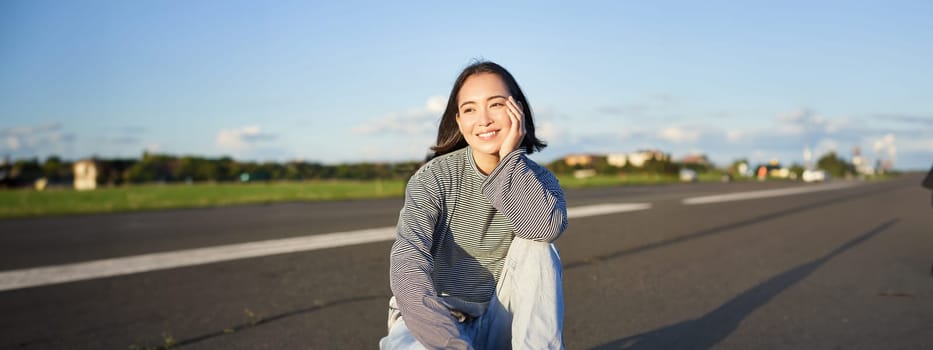 Vertical shot of asian girl skater, sits on her skateboard and smiles, enjoys sunny day, cruising on longboard on empty road outdoors.