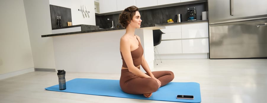 Healthy lifestyle. Young fitness girl does pilates, yoga exercises from home, sits on workout mat in kichen, listens to meditation sounds on mobile phone, uses smartphone gym app.