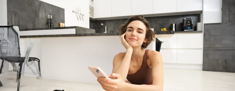 Technology and sport concept. Fitness girl looking at her smartphone, sitting in workout clothes at home, watching exercise videos on mobile phone.