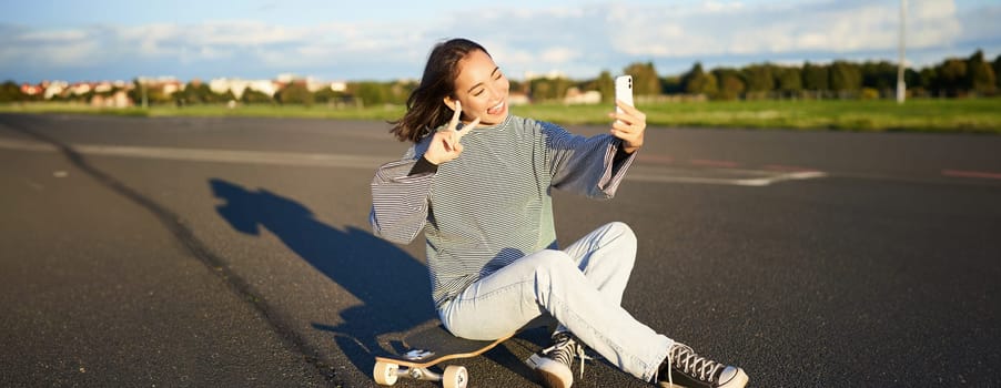 Beautiful korean girl takes selfie on smartphone, takes photo with her skateboard, enoys sunny day outdoors.