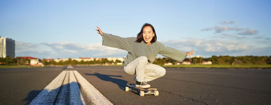 Freedom and happiness. Happy asian girl riding her longboard on an empty sunny road, laughing and smiling, skateboarding.