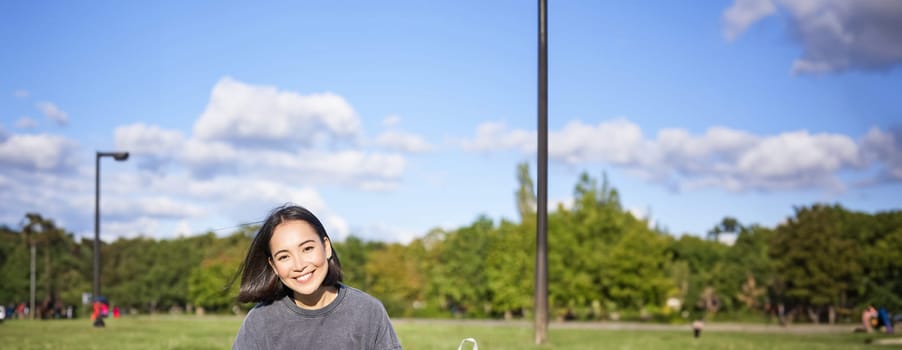 Vertical shot of cute teen girl sits in park on grass with backpack and her book, reading alone outdoors.