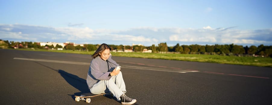 Portrait of young korean girl sitting on her skateboard on road, looking at smartphone, chatting on mobile app.