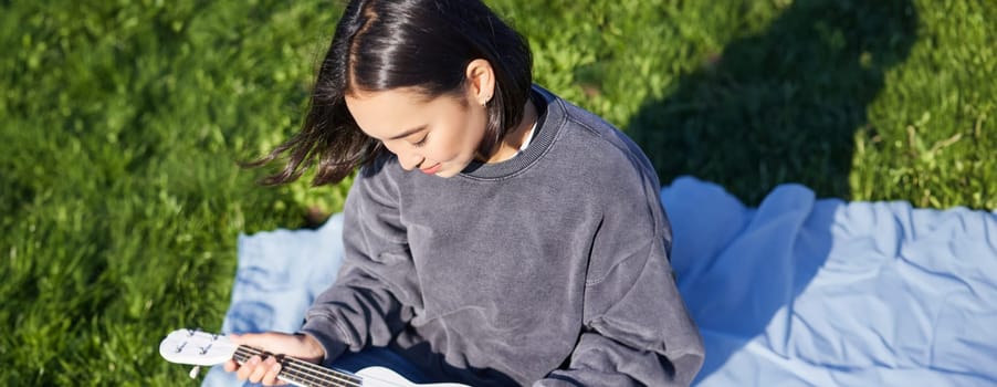 Vertical shot of girl musician, looking with care at her white ukulele guitar, playing music in park, sitting on grass.