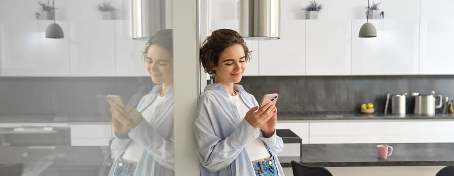 Image of young smiling woman at home, looks at her smartphone with happy face, reads message on telephone, chats on mobile phone app. Technology and communication concept