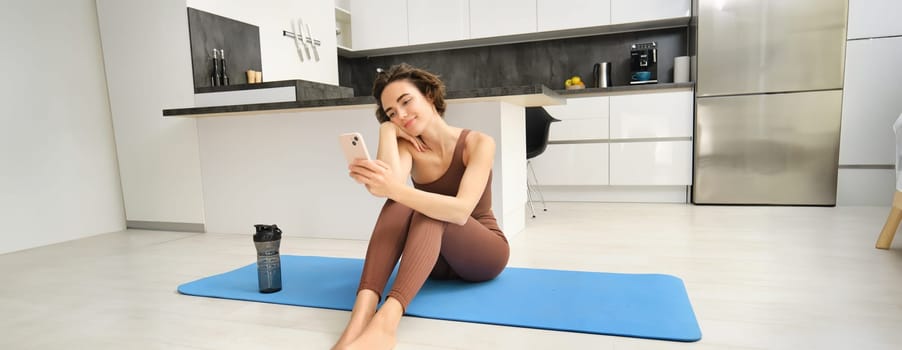 Sport and wellbeing. Portrait of fitness woman, gym instructor workout at home, sits on yoga mat and looks at her smartphone with smiling face.