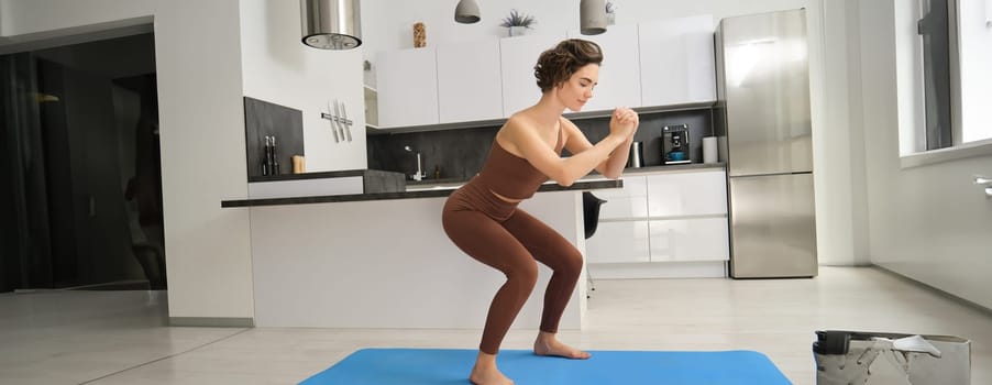 Image of young woman doing squats at home, workout on rubber mat in bright room indoors, wearing activewear for training. Copy space