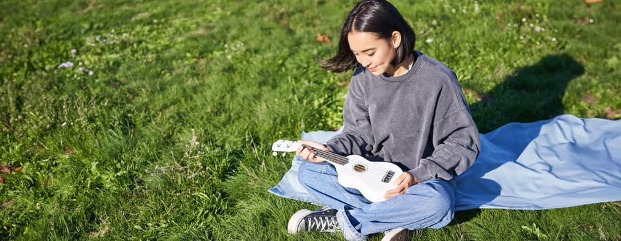 Smiling asian girl, musician playing ukulele in park, looking with care and love at her instrument, sitting on blanket on sunny day.