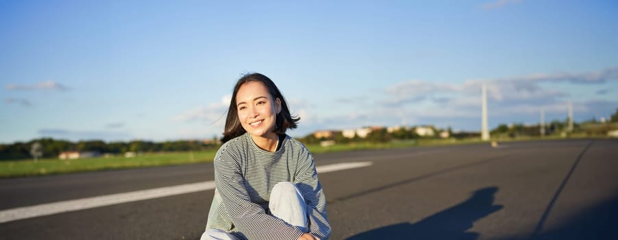 Vertical shot of asian girl skater, sits on her skateboard and smiles, enjoys sunny day, cruising on longboard on empty road outdoors.