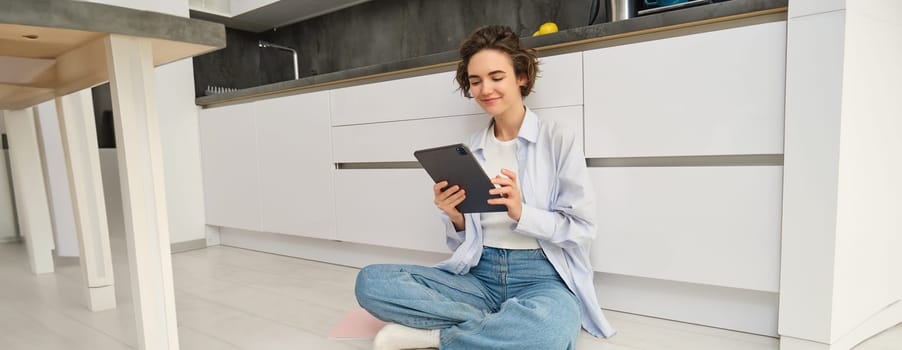 Portrait of young adult woman works on her tablet, sits with notebook on kitchen floor at home, studies in comfort.