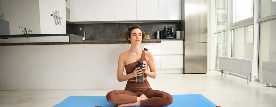 Young strong sporty athletic fitness trainer, instructor woman wearing brown tracksuit sitting on yoga mat drink water training do exercises at home gym indoor. Workout sport motivation concept.