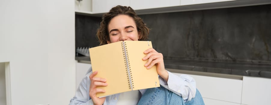 Portrait of candid smiling woman, cover her face with notebook, laughing carefree, doing her homework, studying indoors. Copy space