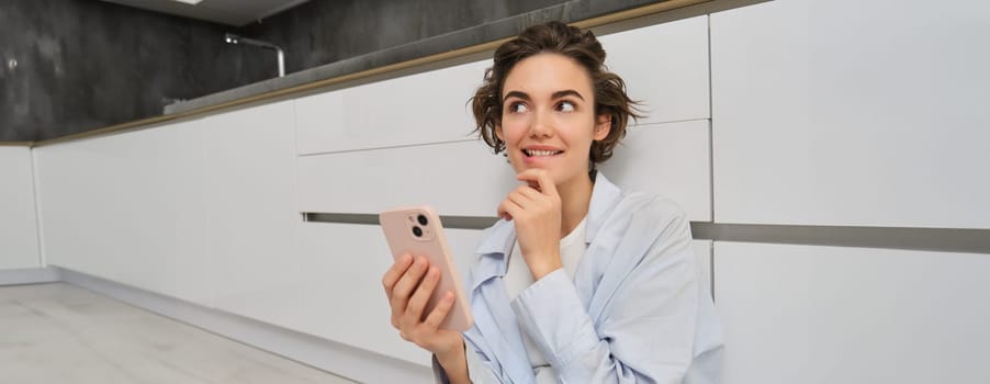 Image of smiling woman thinking, using smartphone and pondering, making decision, online shopping on mobile phone.
