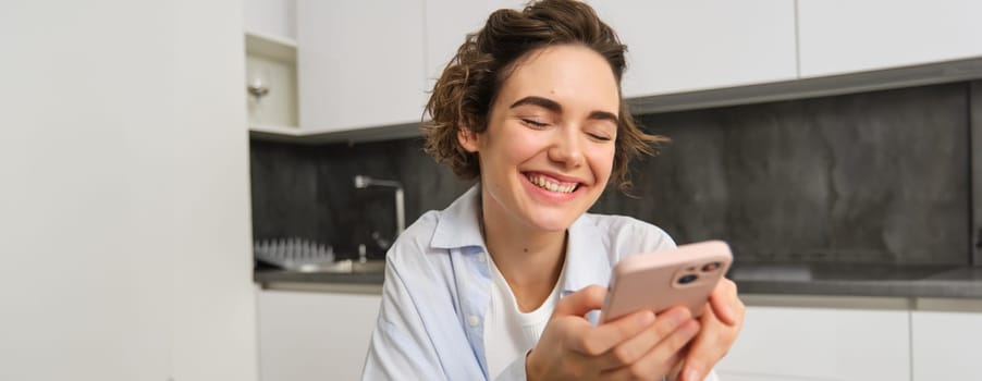 Portrait of modern brunette woman holding mobile phone and smiling. Girl sits in kitchen at home with smartphone, using app to order delivery, does online shopping.
