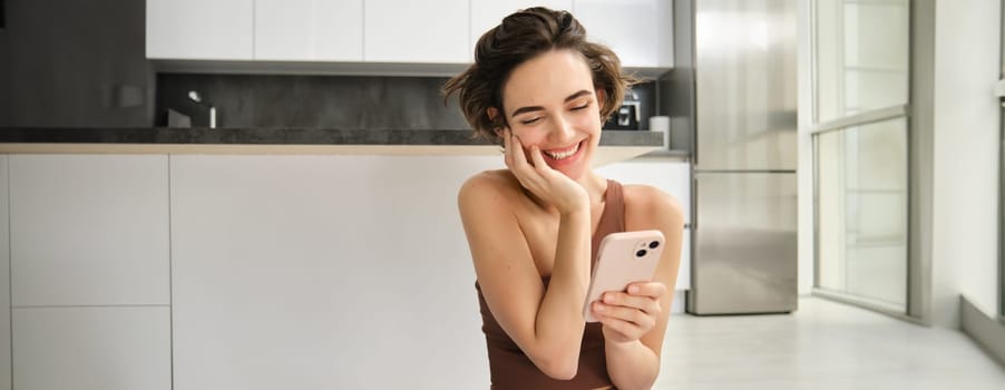Portrait of smiling woman at home, using her mobile phone, sitting on floor in bright room, looking at smartphone and chatting, texting someone. Copy space