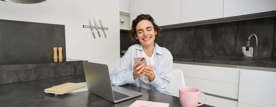 Image of young woman using her smartphone at home. Girl sits with mobile phone in kitchen and smiles.