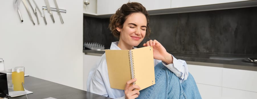 Portrait of carefree brunette woman, laughing and smiling while reading funny text in notebook, reading journal. Copy space