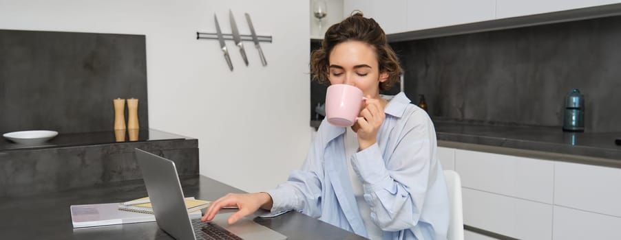 Working woman using laptop, drinking coffee. Girl with cup of tea, studying from hyome, sitting near computer in kitchen, preparing for exam.
