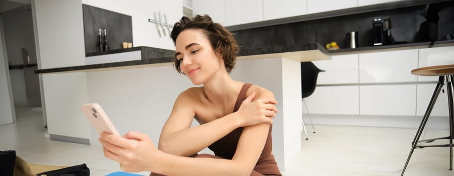 Sport and wellbeing. Portrait of fitness woman, gym instructor workout at home, sits on yoga mat and looks at her smartphone with smiling face.