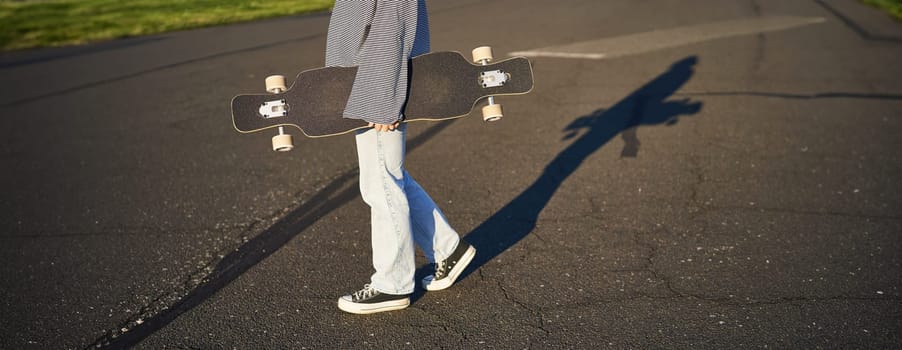 Cropped shot of teen girl body, holding cruiser longboard in hand, walking in sneakers on road in jeans and sweatshirt. Young woman skater with skateboard.