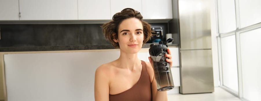 Fitness instructor, smiling young woman workout at home, sits on yoga mat and drinks water, does exercises, training session in her kitchen. Sport indoors concept