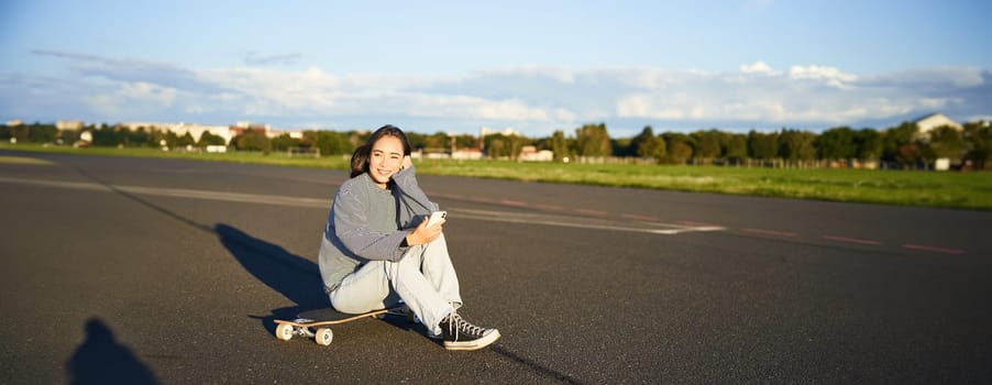 Portrait of young korean girl sitting on her skateboard on road, looking at smartphone, chatting on mobile app.